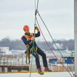 DPI:Scatto dinamico di un lavoratore che esegue un "addestramento uso DPI" per DPI anticaduta , sospeso con imbracatura di sicurezza e cordini. Contesto: cantiere o piattaforma elevata.