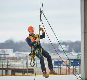 DPI:Scatto dinamico di un lavoratore che esegue un "addestramento uso DPI" per DPI anticaduta , sospeso con imbracatura di sicurezza e cordini. Contesto: cantiere o piattaforma elevata.
