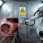 Rischio esplosione: Dettaglio fotografico di una cabina di verniciatura industriale. Un braccio robotico sta spruzzando vernice nebulizzata; si nota la segnaletica gialla "EX" (ATEX) ben visibile sulla parete metallica sullo sfondo. Luci LED fredde.